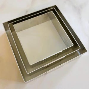 May include: Three square cake pans, nested together. The pans are made of silver metal and have a smooth, shiny finish.