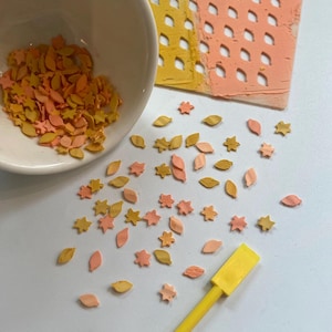 May include: A white bowl filled with small, flat, leaf and star-shaped clay pieces in shades of yellow, orange, and peach. Scattered pieces lie on the surface alongside a yellow plastic tool and a rectangular stencil with a grid pattern.