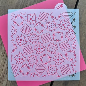 Peut inclure: Un pochoir &agrave; g&acirc;teau violet clair avec un motif r&eacute;p&eacute;titif de carr&eacute;s, de losanges et de points roses. Le pochoir comporte une languette avec la lettre "A". Il est pos&eacute; sur une carte rose, avec une surface en bois en dessous.