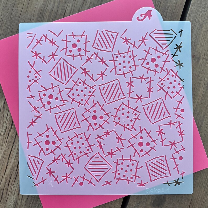 Peut inclure: Un pochoir &agrave; g&acirc;teau violet clair avec un motif r&eacute;p&eacute;titif de carr&eacute;s, de losanges et de points roses. Le pochoir comporte une languette avec la lettre "A". Il est pos&eacute; sur une carte rose, avec une surface en bois en dessous.
