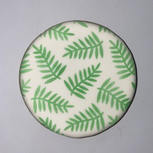May include: A round, white cookie decorated with a pattern of green fern leaves. The leaves are evenly spaced across the surface of the cookie. The cookie has a light brown edge.