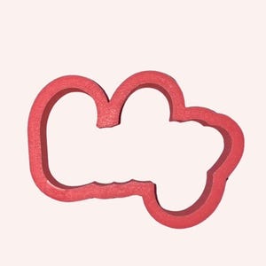 May include: A red cookie cutter with a textured surface, featuring an irregular, rounded, cloud-like shape with three distinct lobes and a hollow centre, designed for cutting dough into a specific outline for baking and food preparation.