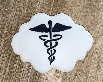 May include: A white, scalloped-edge cookie featuring a black caduceus symbol printed in the center. The medical emblem consists of a central staff with two wings at the top and two snakes coiled around the staff in a symmetrical, intertwined pattern.