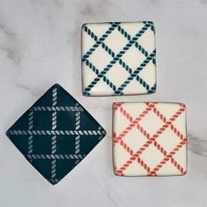 May include: Three square cookies decorated with a rope pattern. The cookies are decorated with blue, teal, and red rope patterns on a white background.