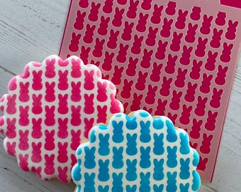 May include: Two cookies decorated with a pattern of pink and blue bunny shapes. A pink stencil with the same bunny pattern is shown in the background.