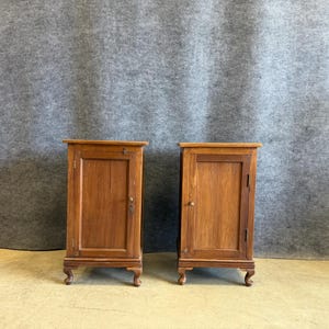 May include: A pair of matching wooden bedside cabinets, each featuring a single rectangular door with a small round knob, a flat top surface, and four curved cabriole legs. The wood has a medium brown finish with visible grain patterns.