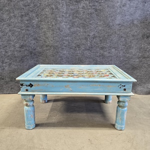 May include: A square, low-profile coffee table with a distressed, light blue painted finish. The tabletop features a mosaic design with various colors and patterns. The table has four sturdy, turned legs, and is suitable for a living room or lounge area.