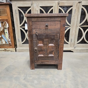 May include: A dark brown wooden end table with a single drawer and a cabinet door with a carved design. The table has a distressed finish and metal hardware.