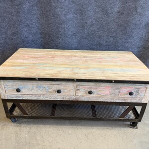 May include: A rectangular coffee table featuring a weathered, multi-colored wood top with hints of blue, pink, and white. The base is constructed from dark metal with two drawers, a lower wire mesh shelf, and four small wheels for mobility.