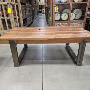 May include: A rectangular wooden coffee table with a natural finish. The tabletop is supported by two angled, dark metal legs. The table is a warm brown color and appears to be made of solid wood. The design is modern and rustic.