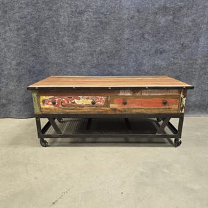 May include: A rectangular coffee table with a wooden top and two drawers. The drawers have a colorful, distressed finish with red, yellow, and green accents. The table sits on a black metal frame with wheels.