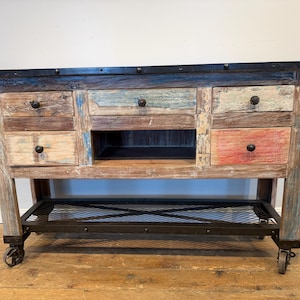 May include: A rectangular wooden kitchen island featuring a distressed, multi-colored finish with blue, red, and natural wood tones. It includes four drawers with black knobs, a central open storage compartment, a black metal top, and a lower mesh shelf.