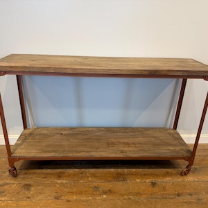 May include: A rectangular console table featuring two horizontal wooden shelves with a weathered brown finish. The structure is supported by a thin, rust-colored metal frame with four small wheels attached to the base of each leg for mobility.