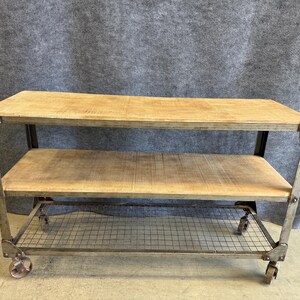 May include: A rectangular rolling cart featuring two light brown wooden shelves and a bottom wire mesh shelf. The frame is constructed from weathered gray metal with visible rivets at the corners and four small wheels attached to the base.