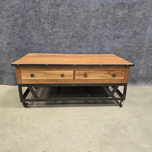 May include: A rectangular coffee table with a wooden top and two drawers. The drawers have black knobs and are set into a wooden frame. The table has a metal base with a shelf and is mounted on wheels.