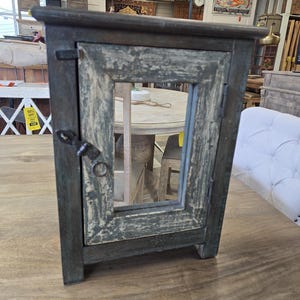 May include: A small, distressed-finish cabinet with a single door and a rectangular mirror. The cabinet is painted in shades of gray and green, with visible wear and tear. It features a metal latch and hinges, and sits on a wooden surface.