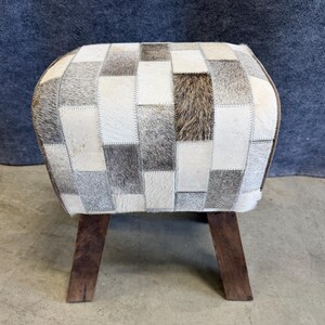May include: A rectangular footstool featuring a patchwork design of cowhide in shades of white, gray, and brown. The top surface is composed of stitched rectangular panels, supported by four dark brown wooden legs with a tapered shape.