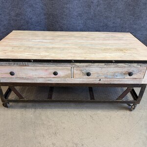 May include: A rectangular coffee table featuring a light-toned, weathered wood top with a dark metal trim. The base includes two drawers with dark round knobs, a lower black metal mesh shelf, and four small wheels attached to the bottom frame.