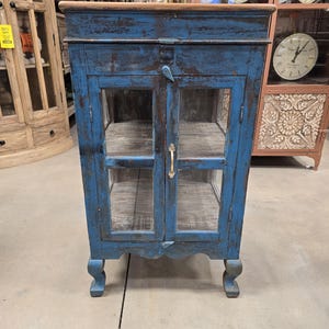 May include: A distressed blue wooden cabinet with glass doors and a light brown top. The cabinet has two shelves inside and a gold-colored handle. The cabinet is supported by four curved legs.