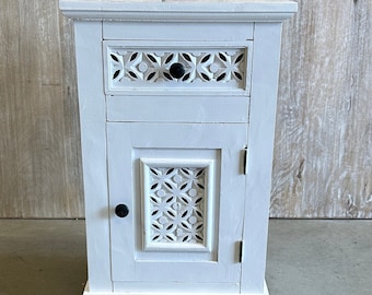 May include: White wooden nightstand with a single drawer and a cabinet door. Both the drawer and door have a carved, geometric design. The nightstand has black knobs.