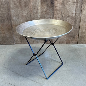 May include: A round, silver tray table with a hammered finish and a black, foldable metal stand.