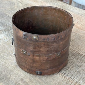 May include: A cylindrical metal container with a dark, weathered brown finish. The vessel features two horizontal metal bands secured by square rivets around the exterior. A small metal ring is attached to the side of the upper band.