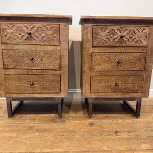May include: A pair of wooden nightstands, each featuring three drawers with intricate carved geometric patterns and small dark metal knobs. The nightstands are supported by dark metal U-shaped legs, providing a sturdy base for the wooden cabinet structure.