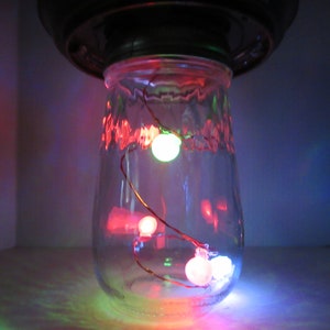 May include: A clear glass jar with a string of colorful LED lights inside. The lights are red, green, and white. The jar is sitting on a white surface.