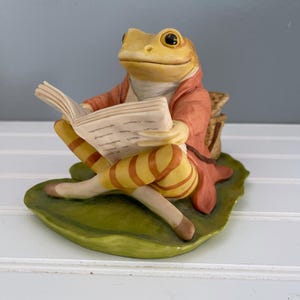 May include: A ceramic frog figurine sitting on a green lily pad, wearing a coral-colored jacket and yellow-striped pants. The frog is holding an open book with visible pages, and a small woven basket is positioned behind its back.