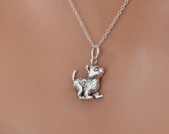 Puede incluir: Un collar de plata con un pequeño dije de plata con la forma de un gatito. El gatito está de pie sobre sus cuatro patas y mirando hacia arriba.