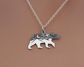 Pode incluir: Um colar com um pingente de urso de prata com um design de montanha e estrela em preto e branco. O pingente está em uma corrente de prata.