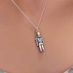 May include: A silver necklace featuring a detailed nutcracker pendant. The pendant is a small, realistic depiction of a nutcracker soldier.