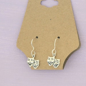 Op de afbeelding: Een paar zilveren oorbellen met een ontwerp van twee theatermaskers, een lachend en een fronsend, hangend aan een bruine kaart.
