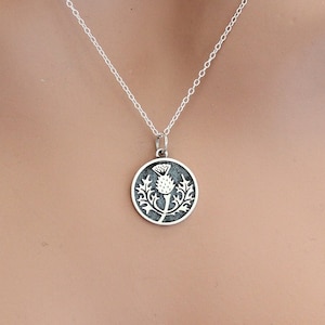 May include: A silver necklace with a round pendant featuring a detailed design of a thistle flower.