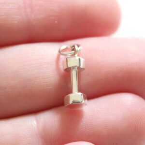 Puede incluir: Un colgante de charm de mancuerna de plata. El charm es pequeño y detallado, con una forma redondeada y un acabado liso.