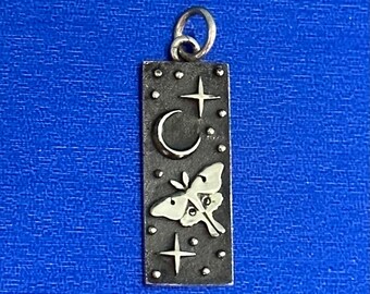 May include: A silver charm with a crescent moon, stars, and a moth design. The charm is rectangular and has a textured surface.
