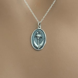 以下が含まれることがあります： 花柄のデザインが施された楕円形のペンダントが付いたシルバーのペンダントネックレス。ペンダントには、葉が付いた花の精巧な画像が描かれています。ネックレスチェーンは繊細でシルバーです。ネックレスは着用されています。