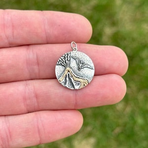 May include: A silver and gold round pendant featuring a detailed depiction of a volcano erupting. The pendant is on a hand, against a green grass background.