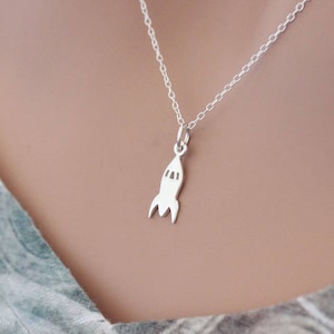 Puede incluir: Un collar de plata con un colgante de cohete. El collar está en un maniquí que lleva una camisa estampada.