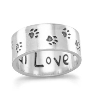 May include: A wide silver-toned band ring featuring a series of small, dark, engraved paw prints around the outer surface. The interior of the ring is inscribed with the word Love in a dark, handwritten-style font, clearly visible from the front.