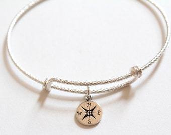 May include: A silver-colored adjustable bangle bracelet with a textured surface. A small, round charm with a compass rose design hangs from the bracelet. The compass rose has the letters N, E, S, and W.