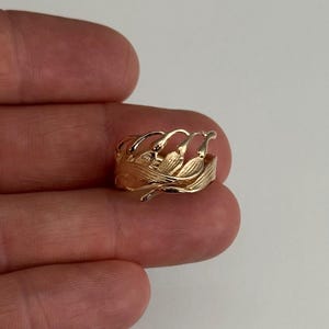 May include: A gold-colored ring with a floral design. The ring features a series of curved, leaf-like shapes that encircle the band. The ring is held in a person's hand.