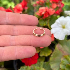 Puede incluir: Un anillo de dedo del pie de oro con un diseño texturizado. El anillo se muestra en un dedo contra un fondo de follaje verde y flores rojas.