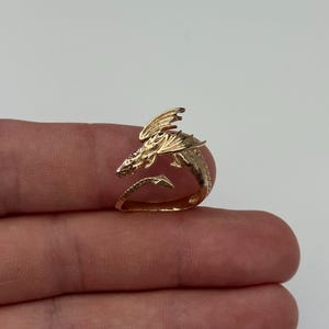 Puede incluir: Un anillo de dragón dorado. El anillo presenta un diseño detallado de dragón con alas y escamas. La cabeza del dragón está colocada en la parte superior del anillo, con la cola envolviendo el dedo. El anillo se muestra en un dedo sobre un fondo neutro.