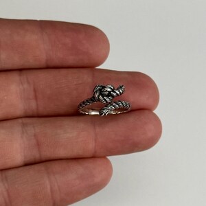 May include: A silver-colored ring in the shape of a nautical knot. The ring has a textured, rope-like design and wraps around the finger. The knot detail is prominent, adding a unique, maritime-inspired aesthetic to the jewelry.