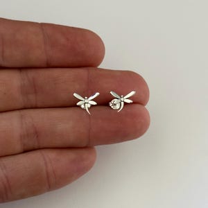 Op de afbeelding: Een paar kleine zilveren oorstekers in de vorm van libellen. Elke oorbel heeft een gedetailleerd libellenontwerp met delicate vleugels en een gebogen staart. De oorbellen worden getoond tegen een neutrale achtergrond.
