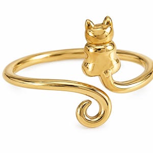 May include: A gold-toned open ring featuring a stylized cat figure sitting upright on one end of the band, with the cat's tail curving into a spiral shape at the opposite end of the open design, creating a continuous smooth metallic surface.