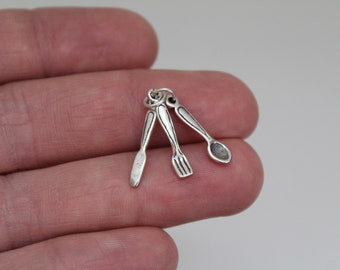 Może przedstawiać: Srebrny charm przedstawiający nóż, widelec i łyżkę. Charm jest zaprojektowany tak, aby wyglądał jak miniaturowe sztućce.