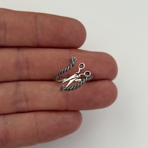 Pode incluir: Um anel prateado com um par de tesouras no centro, rodeado por um design em forma de corda. As tesouras têm lâminas abertas. O anel foi concebido para envolver o dedo, criando um acessório único e elegante.