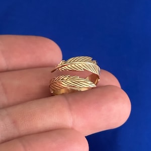 May include: A gold-toned ring with a double feather design. The ring is adjustable and open on one side.